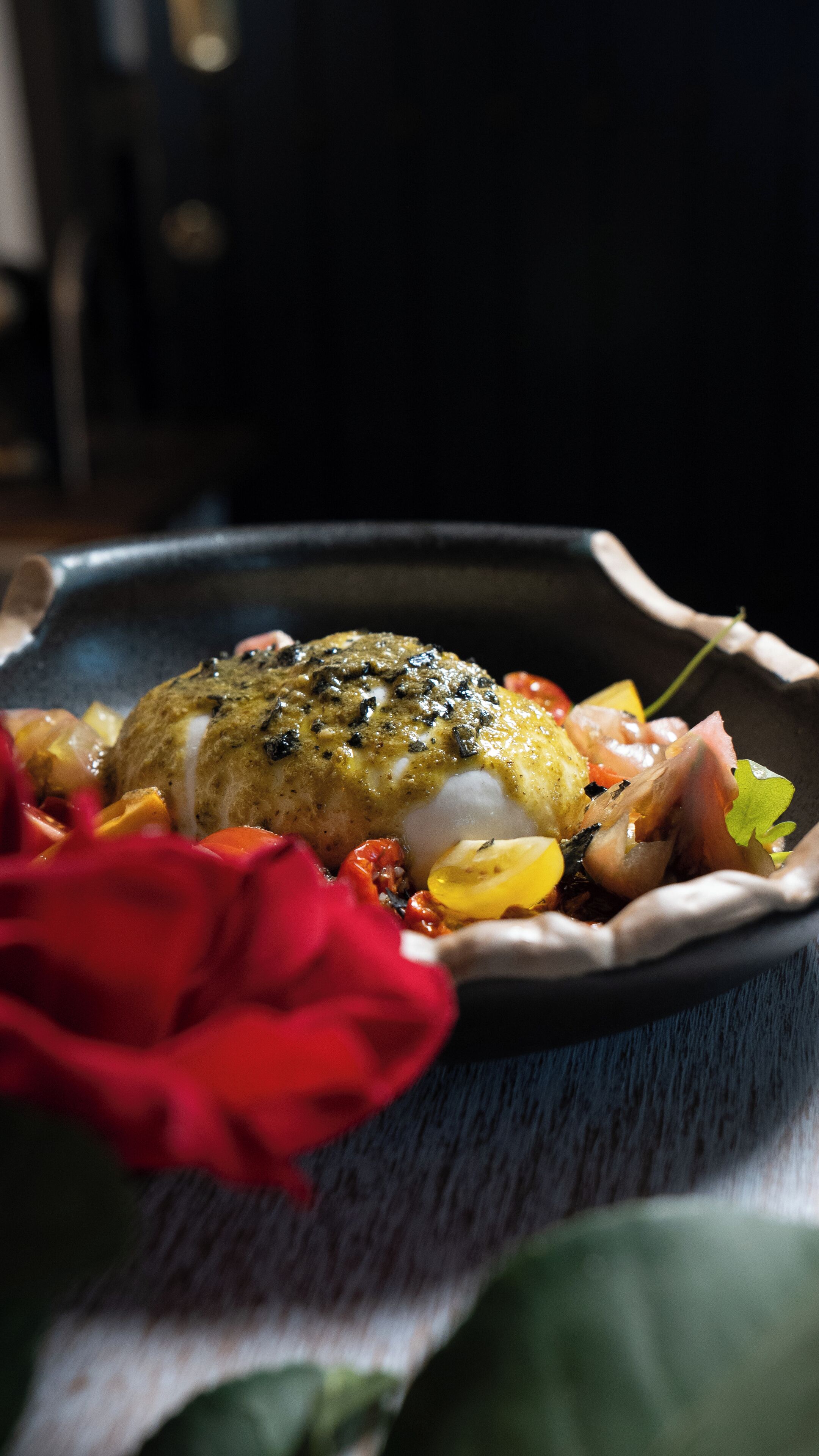A dish with a green herb topping, surrounded by vegetables and garnished with a red flower.