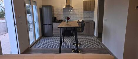 Une cuisine compacte équipée d’un réfrigérateur, d’une cuisinière et d’un évier. On y trouve une table à manger avec deux chaises. La pièce est dotée d’un sol carrelé et d’une porte-fenêtre coulissante donnant sur l’extérieur.