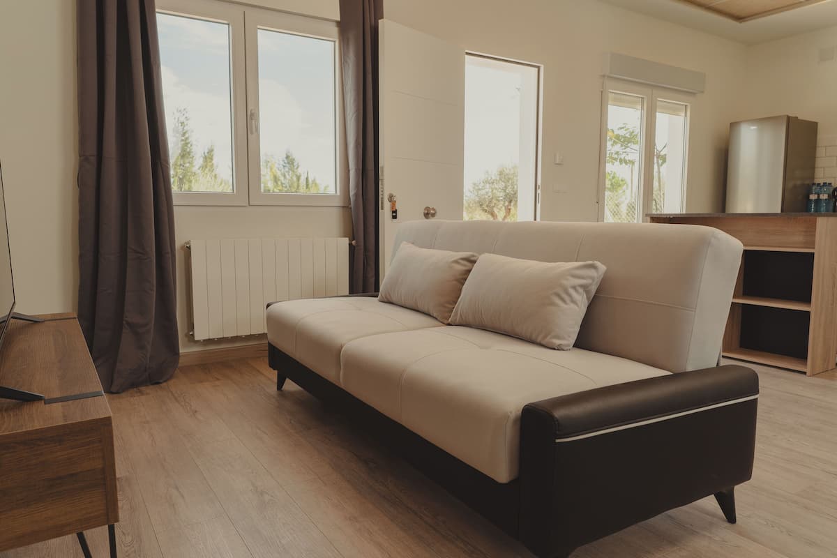 A modern living room with a beige sofa, wooden flooring, a wooden side table, and a kitchen area with a refrigerator and cabinets.