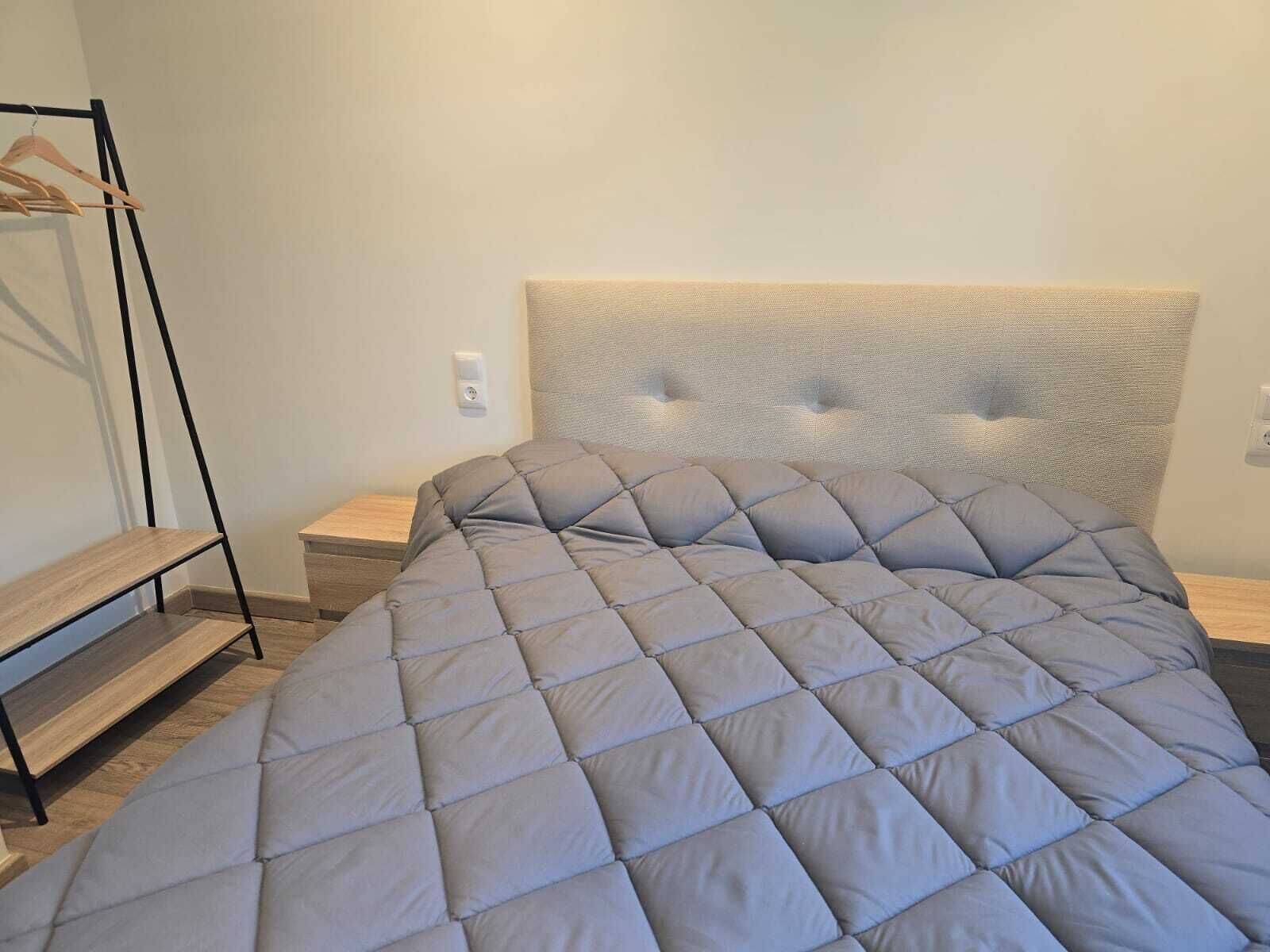 Una cama bien hecha con un colcha gris, una mesita de noche de madera y un perchero metálico negro.
