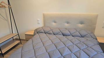 Una cama bien hecha con un colcha gris, una mesita de noche de madera y un perchero metálico negro.