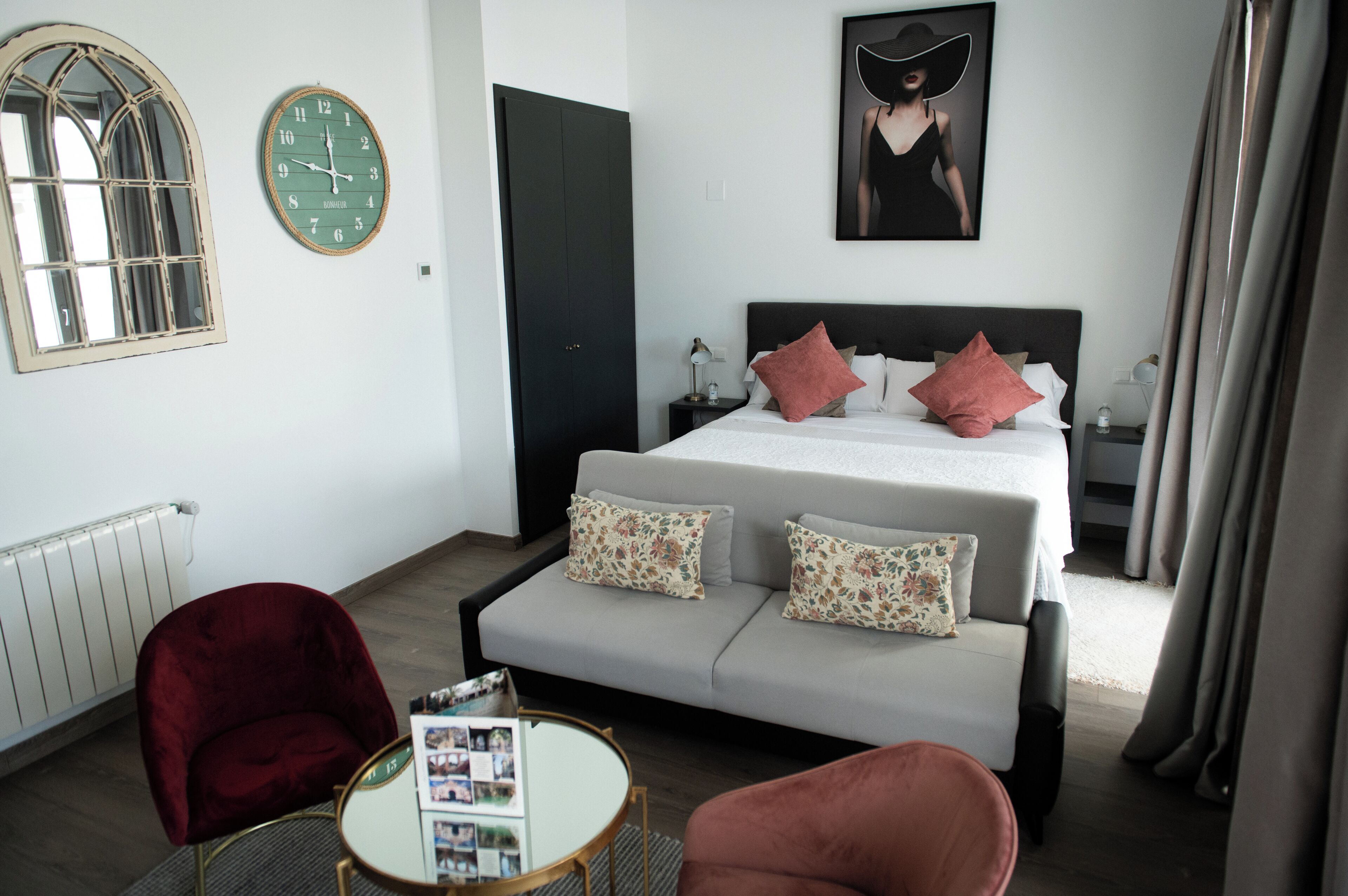 A bedroom with a bed, two nightstands, a round table with magazines, a red chair, and a large mirror.