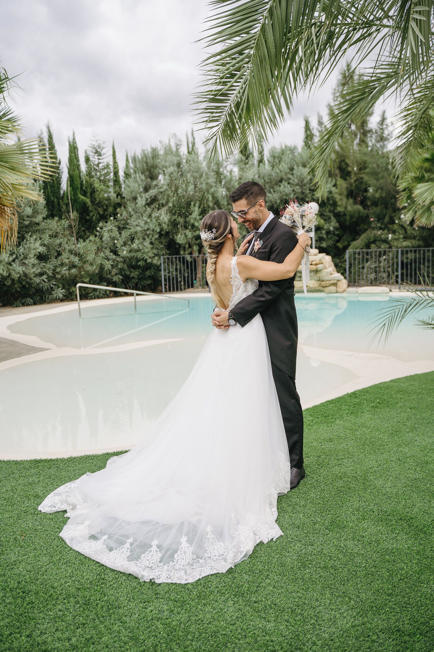 A couple embracing near a pool with a lush green lawn and tropical plants in the background.