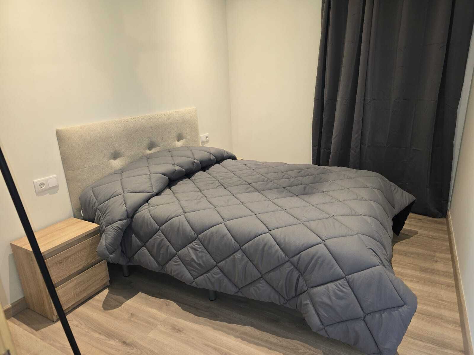 A neatly made bed with a grey quilt in a modern room with wooden flooring and a headboard.