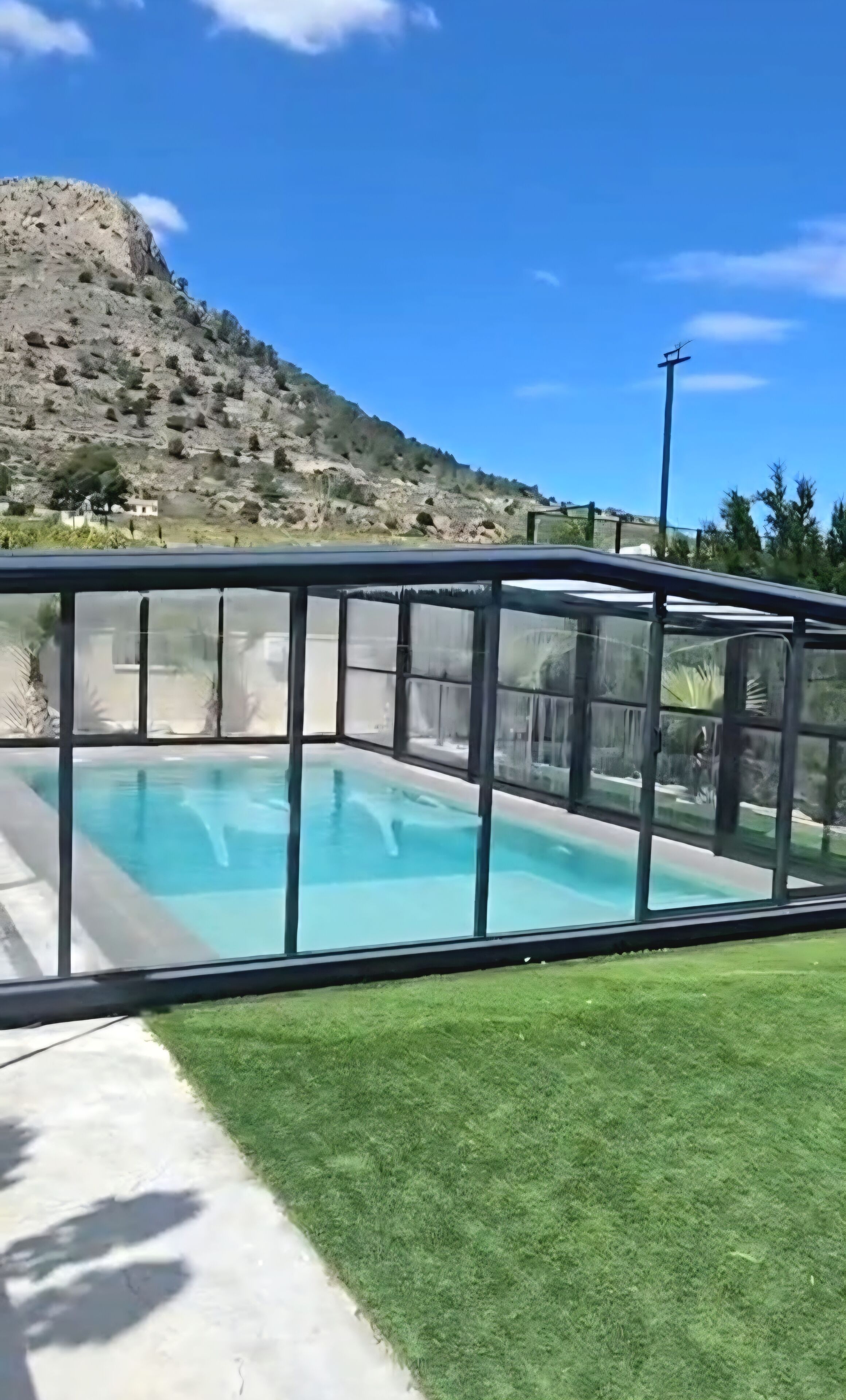 Una piscina con cerramiento de vidrio, rodeada de césped y con una montaña al fondo.