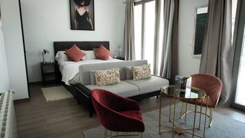 Una habitación de hotel con cama, dos mesitas de noche, una silla roja, una mesita pequeña y una ventana con cortinas.