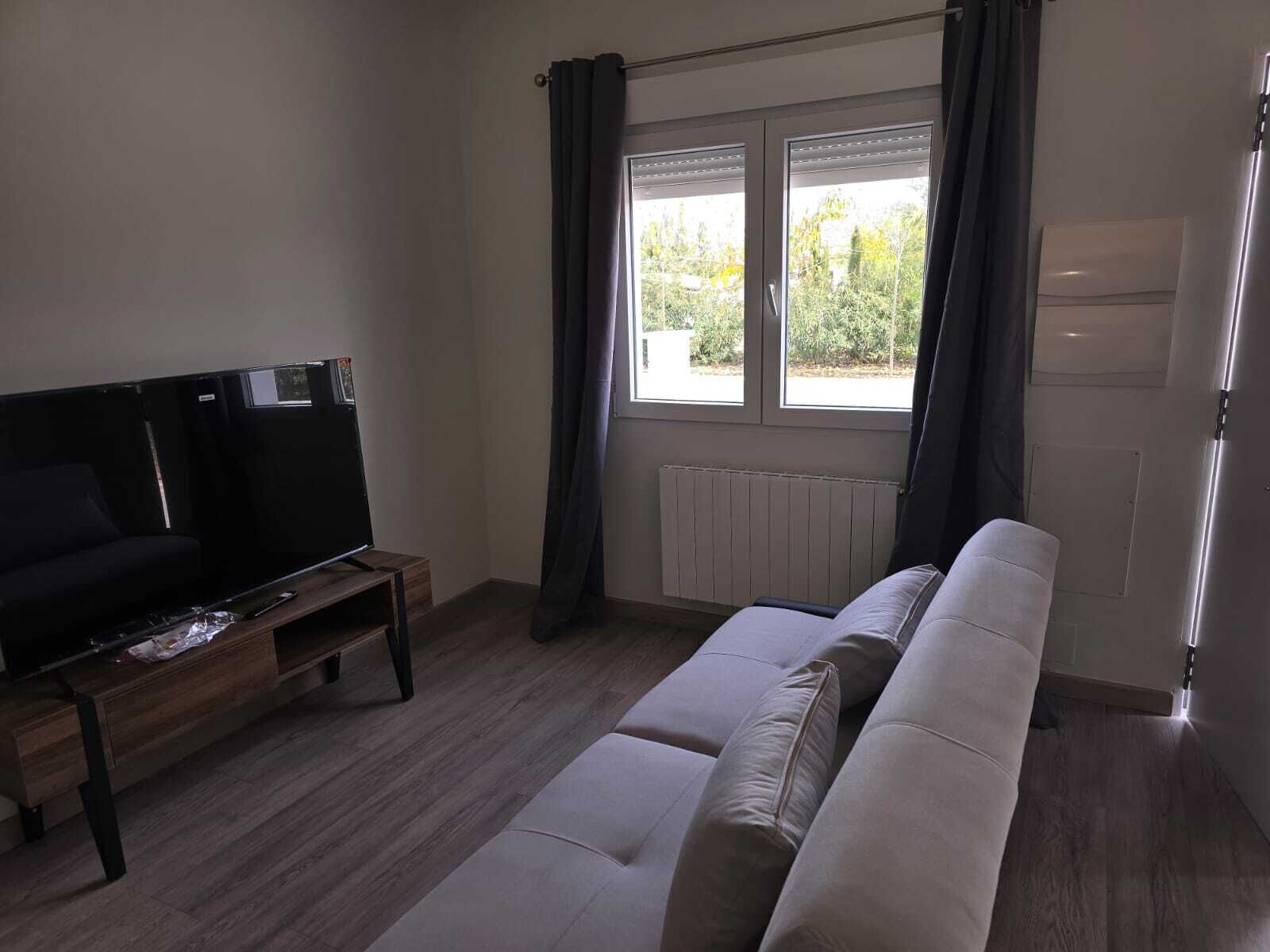 Uma sala de estar com sofá, televisão sobre um suporte de madeira, janela com cortinas e radiador.
