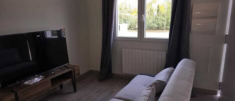 Uma sala de estar com sofá, televisão sobre um suporte de madeira, janela com cortinas e radiador.