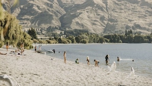 A beach with people and a boat, surrounded by mountains and trees. - Private Balcony, 2 Bathroom, Free Washer Dryer (Wānaka)