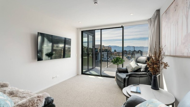 A modern living room with a large sliding glass door leading to a balcony, a mounted TV, and a cozy seating area with a coffee table. - Private Balcony, 2 Bathroom, Free Washer Dryer (Wānaka)