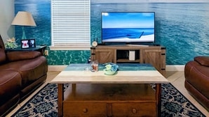 Ett vardagsrum med en stor TV, ett fönster med persienner, ett soffbord och en soffa.