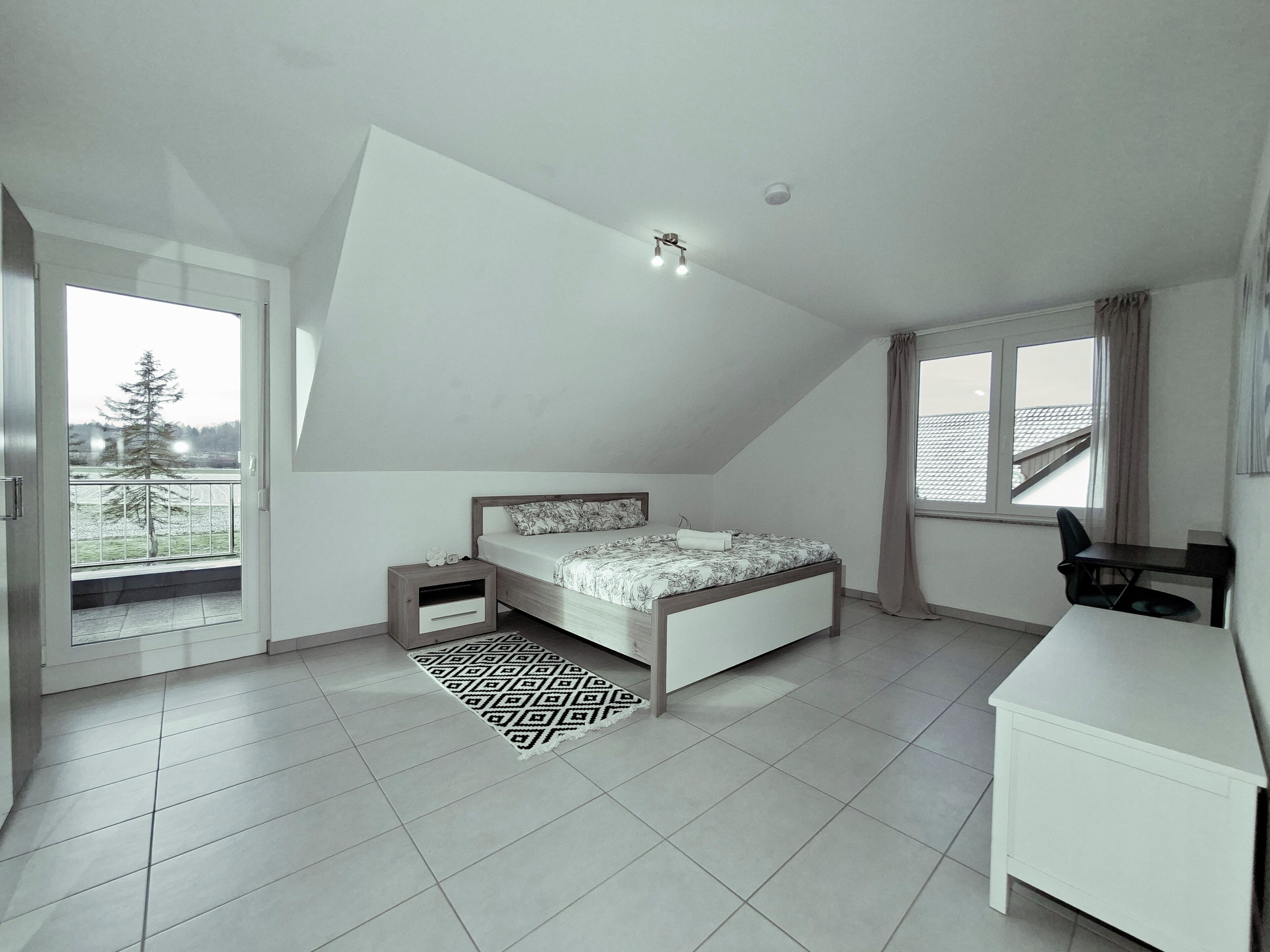 A bedroom with a bed, a desk, a chair, a window, and a balcony.