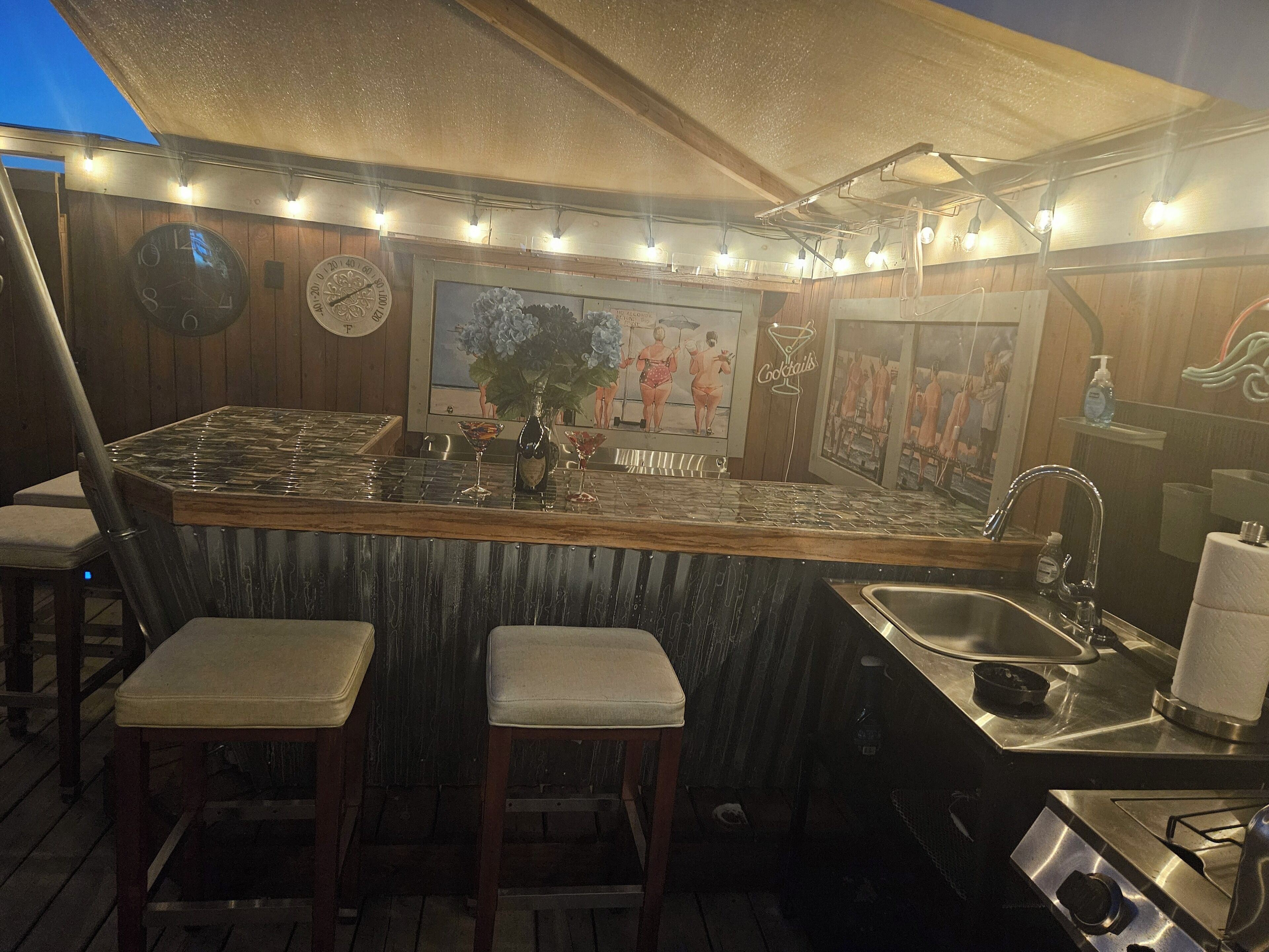 Una zona de bar con paredes de madera, un reloj y un cuadro de personas. Hay dos taburetes y un lavamanos.