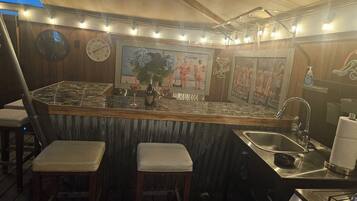 Una zona de bar con paredes de madera, un reloj y un cuadro de personas. Hay dos taburetes y un lavamanos.