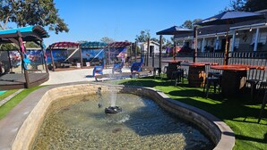 A fountain in a landscaped area with a pool, surrounded by grass and a paved walkway. - Liberty Hill Executive Inn (Zephyrhills)