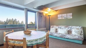 A cozy room with a round wooden table, a sofa, and a view of trees through a large window. - Apartment with Ski Access (Chamrousse)