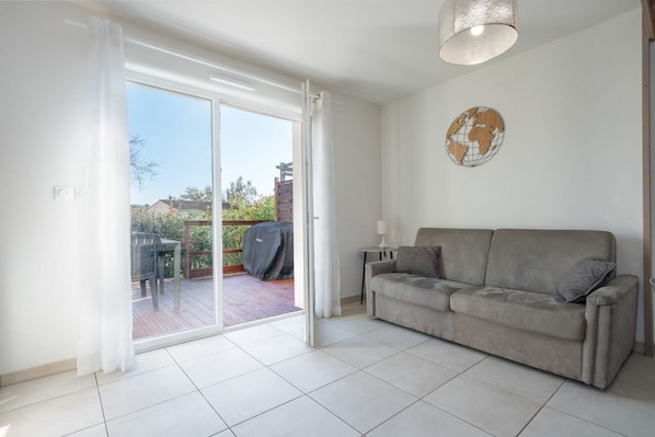 Un salon avec un canapé gris, une table avec une lampe, un globe décoratif et une porte-fenêtre coulissante donnant sur une terrasse extérieure.