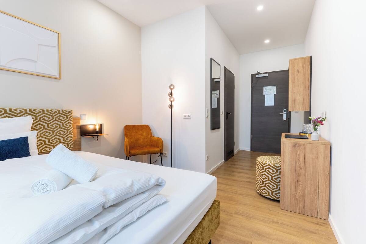 Een moderne hotelkamer met een netjes opgemaakt bed, een houten nachtkastje, een klein poufje, een stoel en een garderobe.