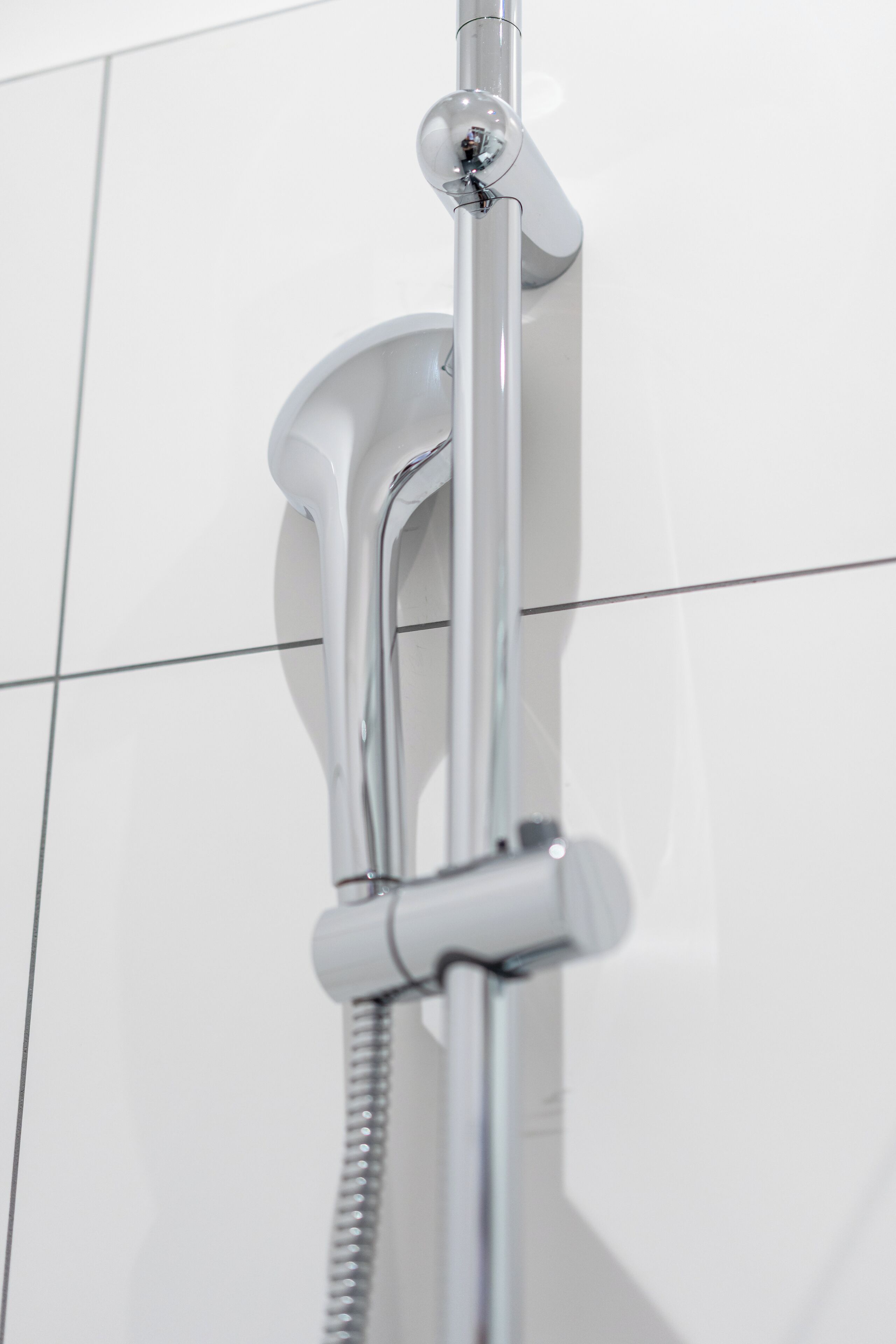 Un pommeau de douche chromé, muni d’un flexible, est fixé sur un mur recouvert de carreaux blancs.