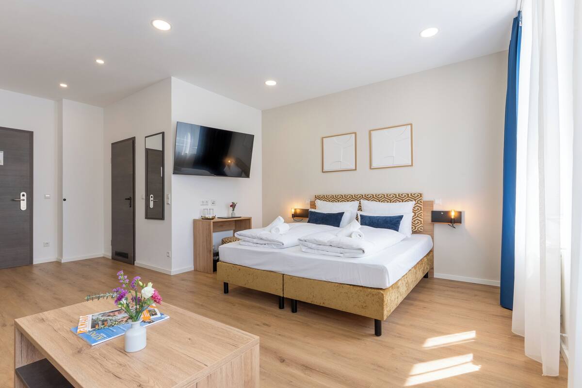 Een moderne hotelkamer met een groot bed, een flatscreen televisie aan de muur, een nachtkastje met een lamp en een houten salontafel met een tijdschrift en bloemen.