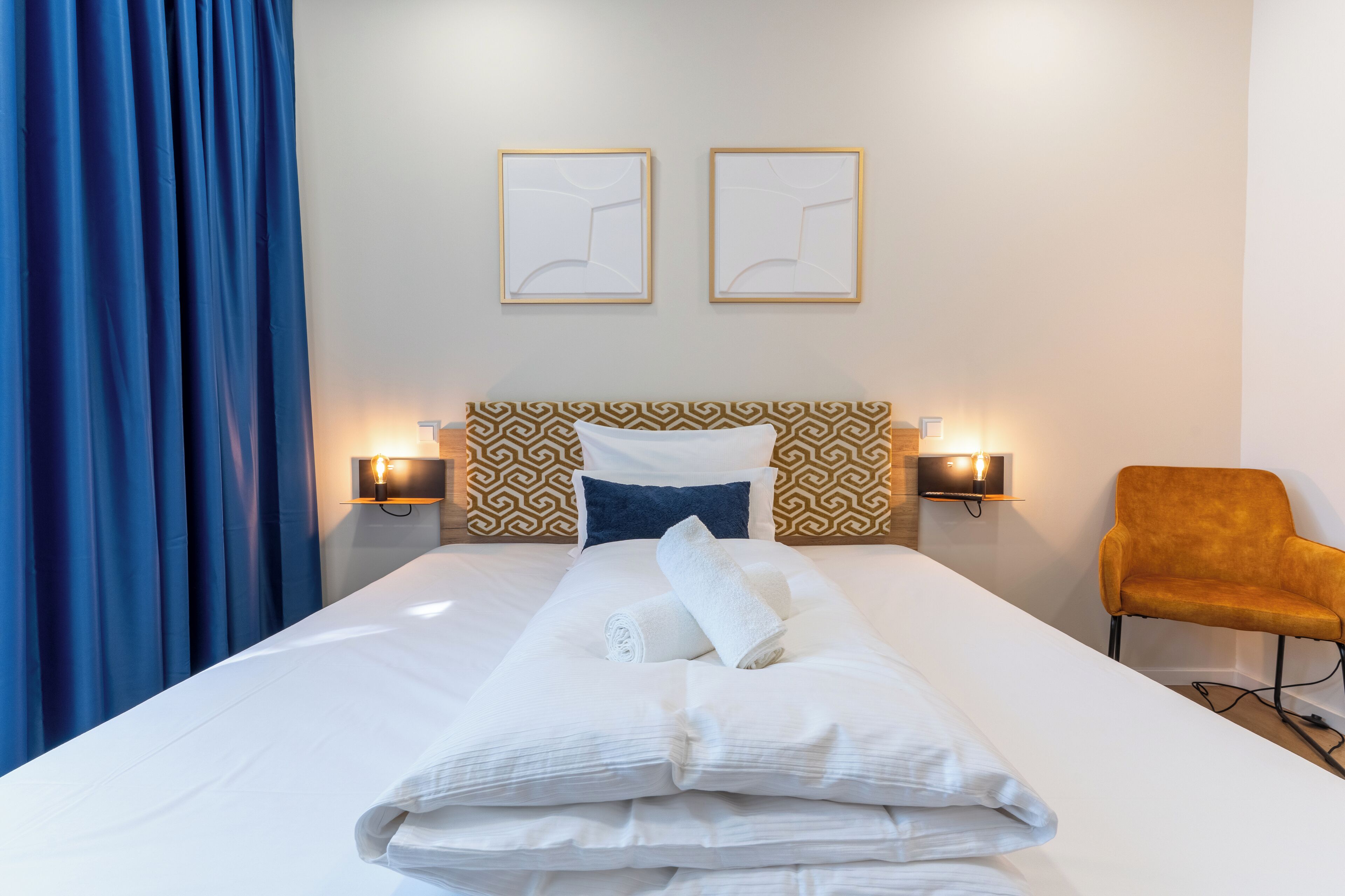 A hotel room with a neatly made bed, two wall-mounted lamps, a patterned headboard, and a single chair.