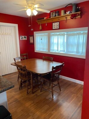 Un coin repas avec une table et des chaises en bois, un ventilateur de plafond, une fenêtre avec des rideaux et un mur rouge.