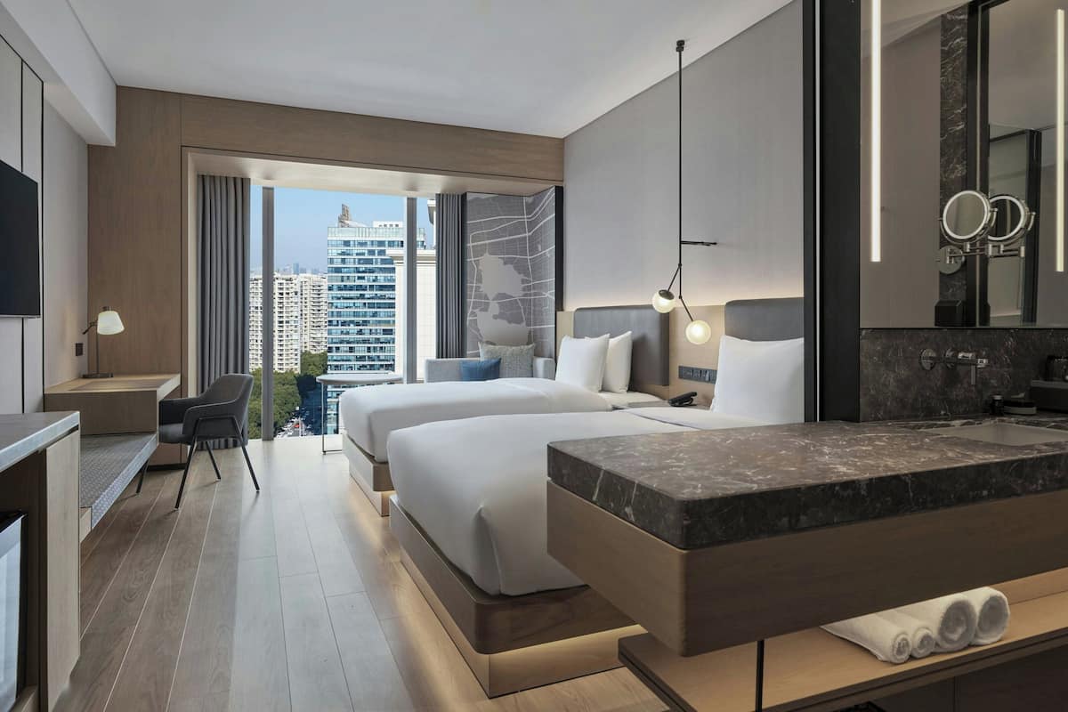 Une chambre d’hôtel moderne avec deux lits, une grande fenêtre donnant sur la ville, un meuble-lavabo en marbre dans la salle de bain et un espace bureau.
