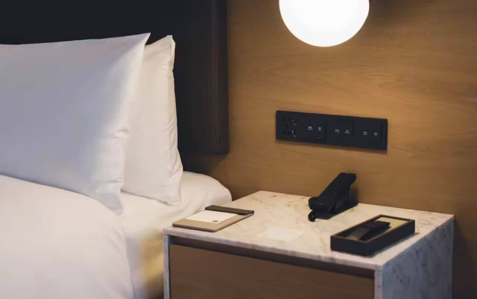Un lit bien fait, avec du linge de lit blanc, une table de chevet avec une lampe, un téléphone et une tablette.