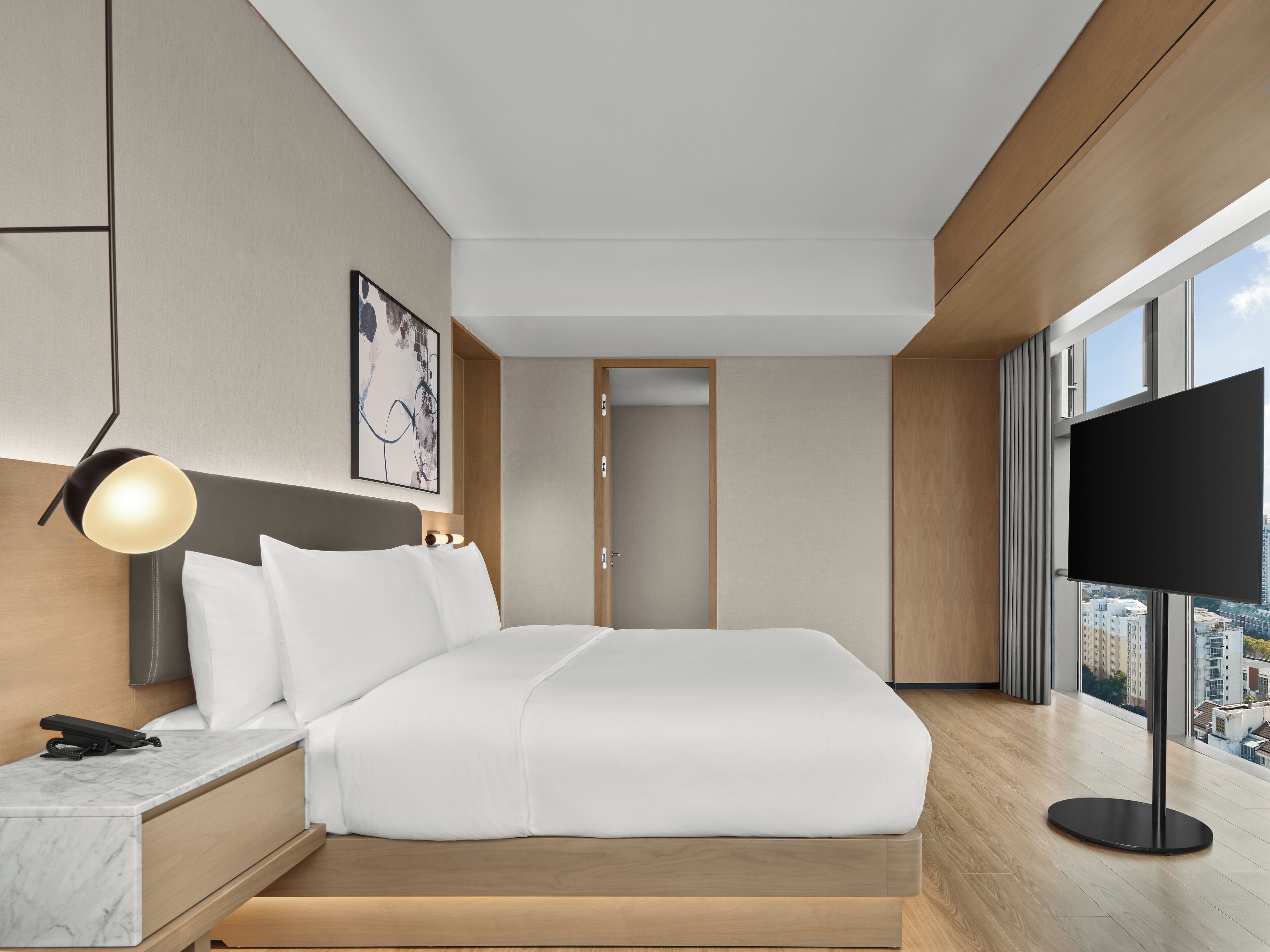 Moderni hotellihuone, jossa on suuri sänky, yöpöytä lampun kanssa, taulutelevisio ja suuret ikkunat, joista avautuu näkymä kaupunkiin.