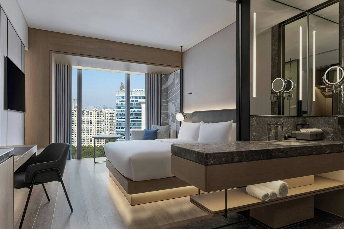 Une chambre d’hôtel moderne dotée d’un grand lit, d’un coin salon, d’une salle de bain avec un grand miroir et offrant une vue sur le paysage urbain.