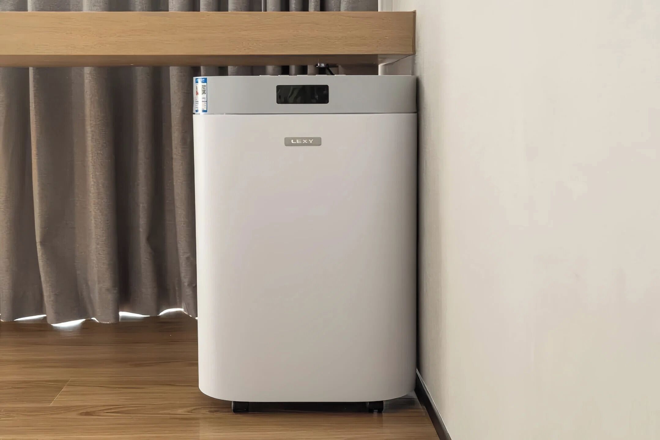 A white, modern, compact refrigerator with a control panel on top, placed against a wall with a wooden shelf above it.