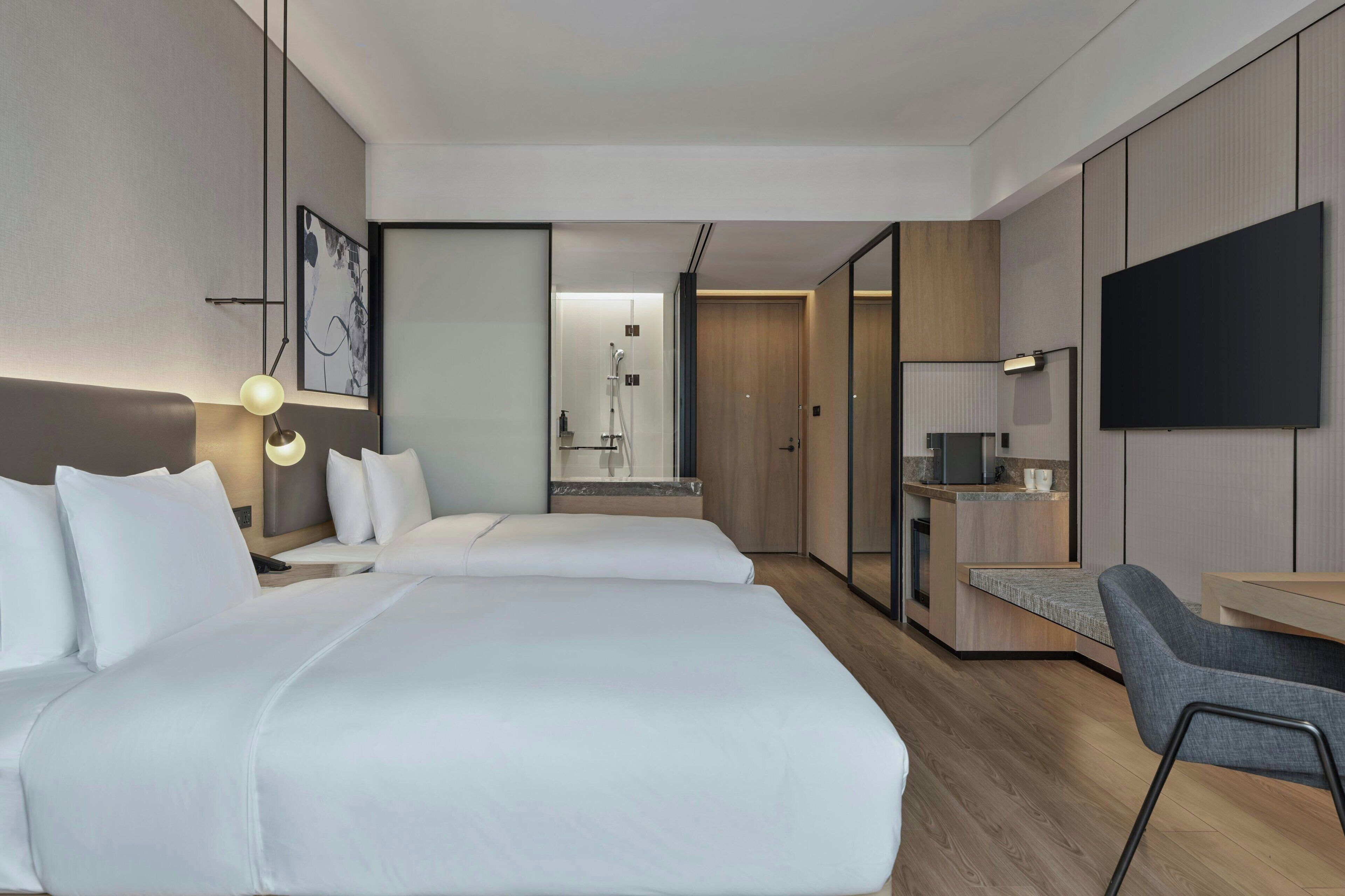 A modern hotel room with two beds, a desk, a chair, a TV, and a shower area.
