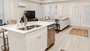 Eine moderne Küche mit Kochinsel, ein Essbereich mit Tisch und Stühlen, ein Wohnzimmer mit Sofa und ein großes Fenster mit Vorhängen.