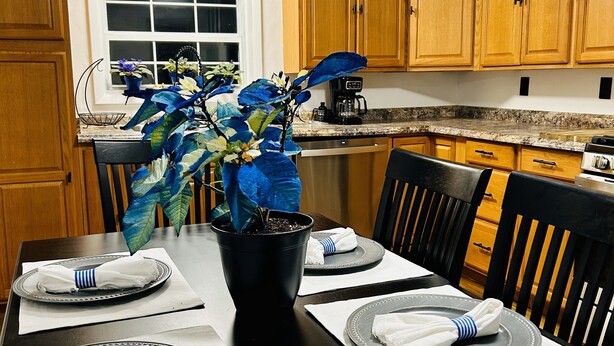 A dining table set for four with a black vase centerpiece, wooden cabinets, and a window above the sink.