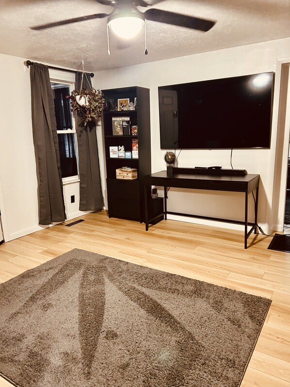 Una habitación con ventilador de techo, televisión de pantalla plana, estantería y una alfombra grande sobre piso de madera.