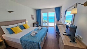 Ein Hotelzimmer mit einem Bett, einem Schreibtisch mit Fernseher, einem Balkon mit Meerblick und einem Fenster mit Vorhängen.