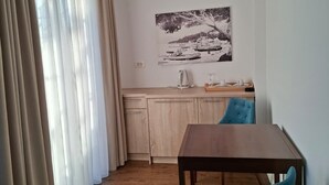 Ein Zimmer mit einem Holzespeisetisch, zwei Stühlen, einer Küchenzeile mit Spüle und Herd sowie einem großen Fenster mit durchsichtigen Vorhängen.