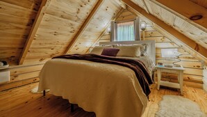 A cozy bedroom with a sloped ceiling, a bed with a patterned bedspread, a nightstand with a lamp, and a window with curtains. -  Luxury in the Smokey Mountains with Hot Tub Pet Friendly (Elizabethton)