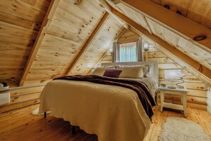 A cozy bedroom with a sloped ceiling, a bed with a patterned bedspread, a nightstand with a lamp, and a window with curtains.