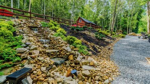 A forested area with a wooden cabin, a small waterfall, and a gravel path. -  Luxury in the Smokey Mountains with Hot Tub Pet Friendly (Elizabethton)