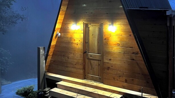 Une cabane en bois avec un toit triangulaire, une porte d’entrée et deux lampes extérieures.