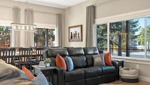 Un salon moderne avec un canapé en cuir noir, une table à manger avec des chaises, un lustre et une grande fenêtre donnant sur des arbres et une rue.