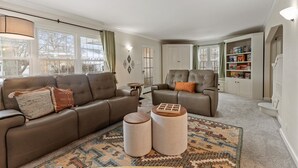 A spacious living room with a brown leather sofa, two ottomans, a patterned rug, and a bookshelf filled with board games.