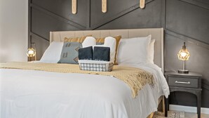 A neatly made bed with a white comforter, two decorative pillows, a small tray with towels, and a bedside lamp on a dark nightstand.