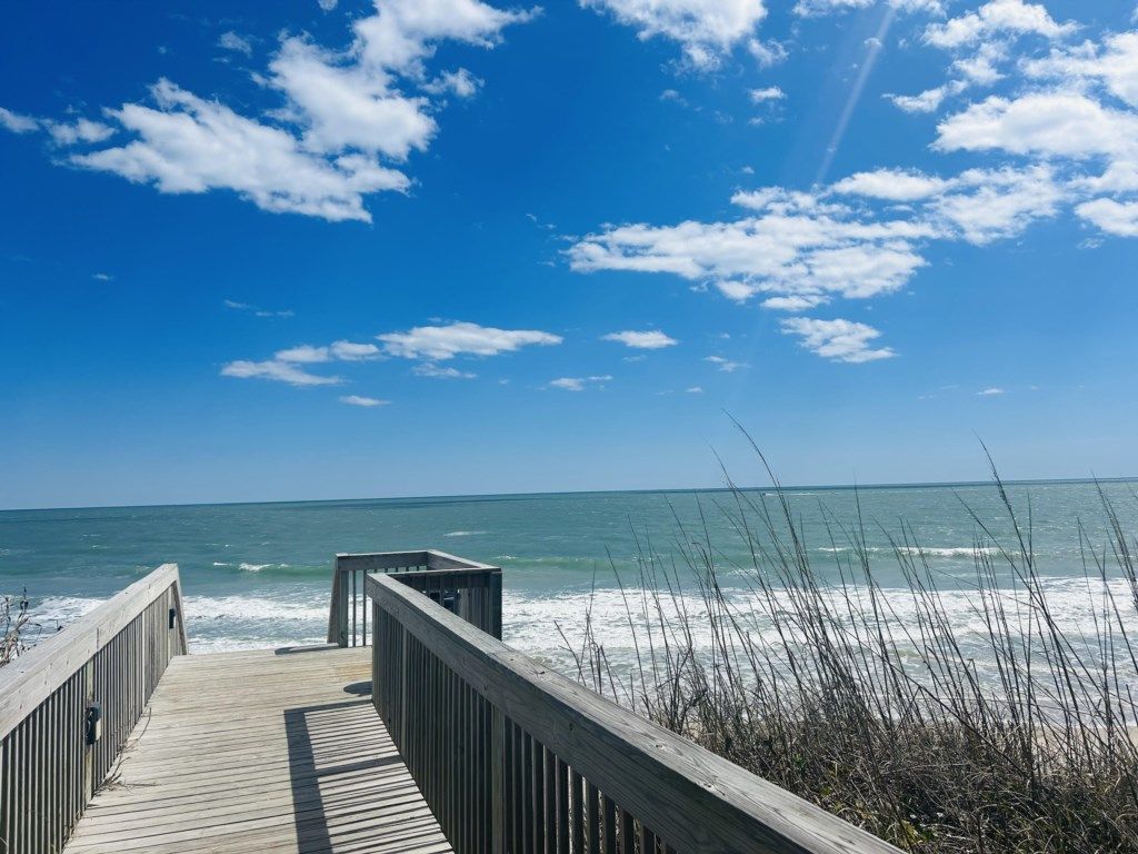 A wooden boardwalk leading to a beach with waves, clear sky, and sparse vegetation.