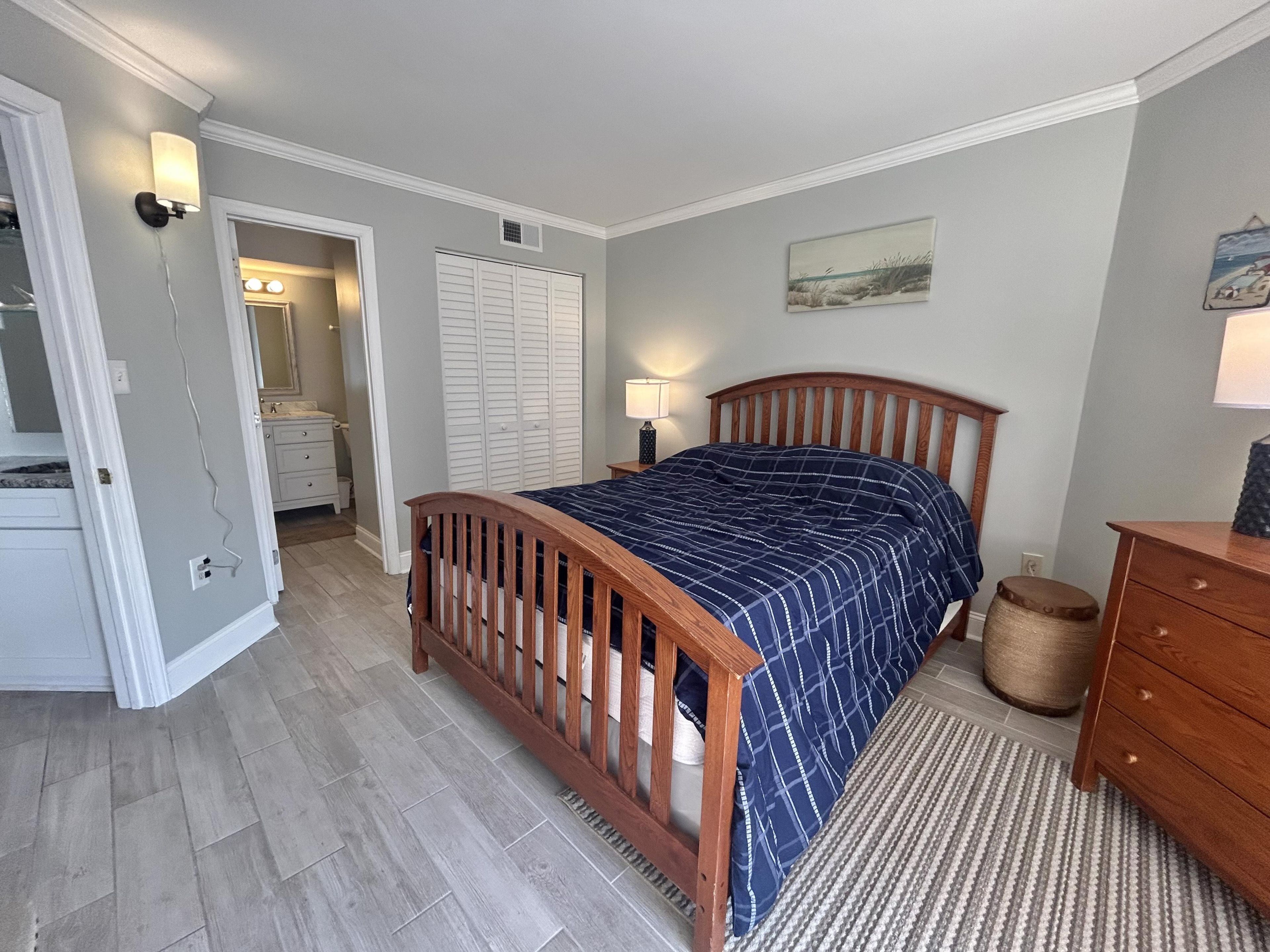 Quarto com cama de madeira, mesa de cabeceira, cômoda, armário e banheiro visível por uma porta aberta.
