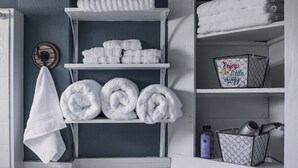 Um banheiro com porta-toalhas, prateleiras com toalhas dobradas e um cesto com artigos de higiene pessoal, além de uma placa decorativa.