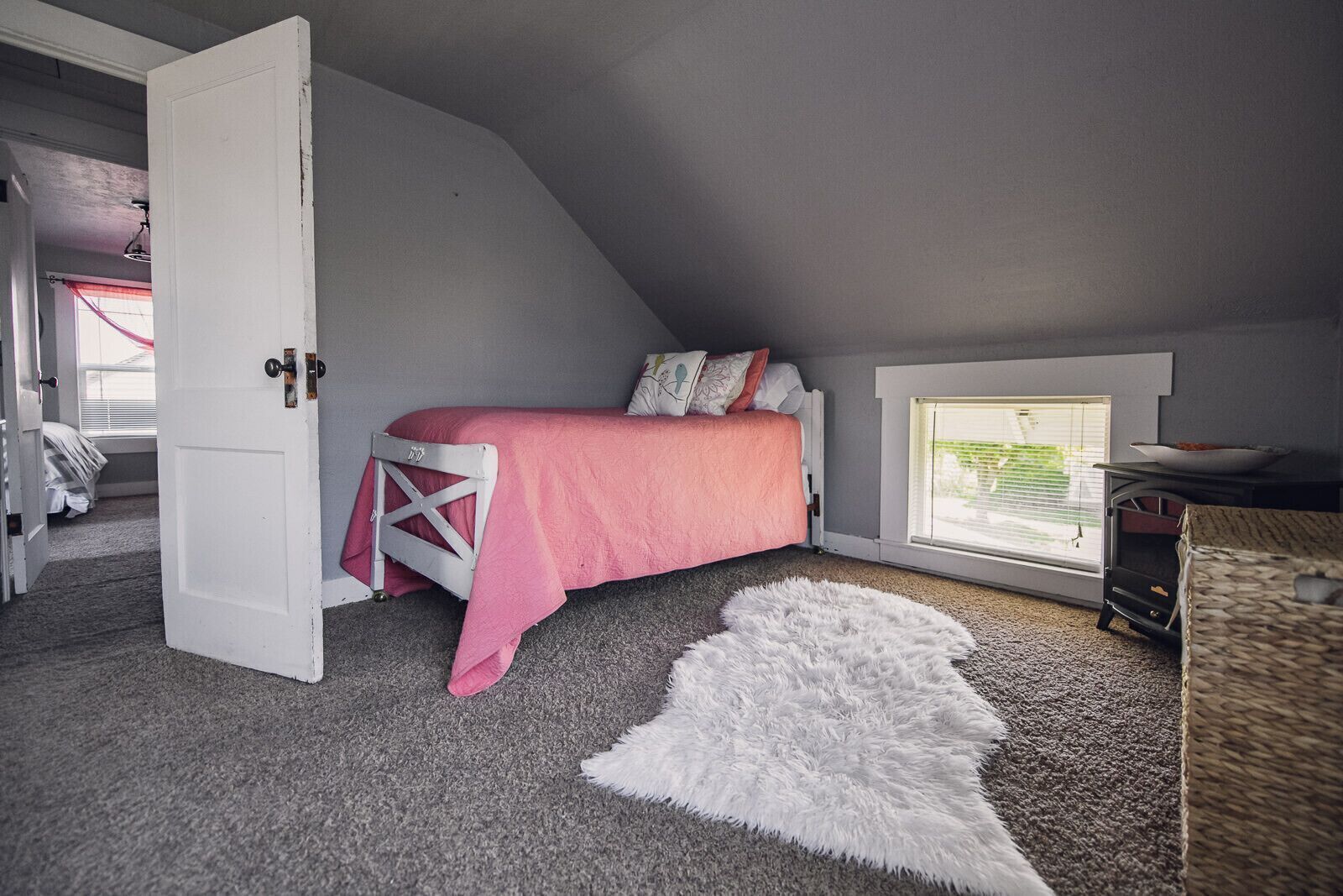 A bedroom with a sloped ceiling, a bed with a pink blanket, a white fluffy rug, and a window with blinds.