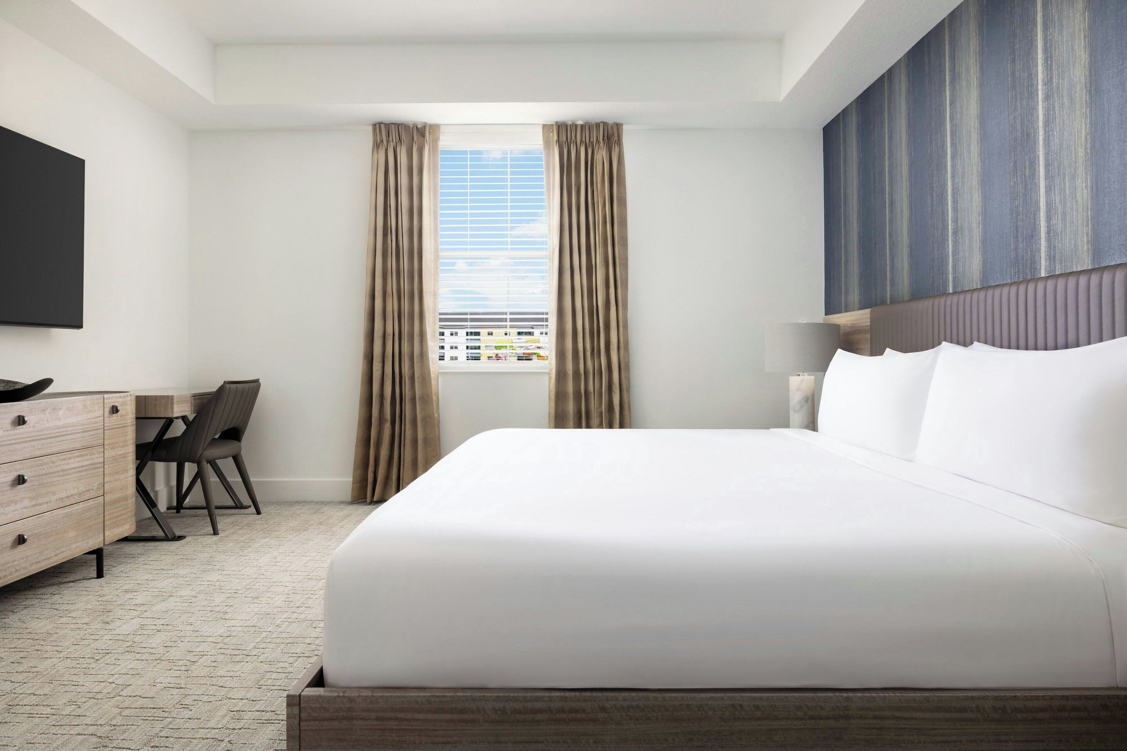 Ein Hotelzimmer mit einem großen Bett, einem Schreibtisch mit Stuhl, einem Flachbildfernseher und einem Fenster mit Vorhängen.