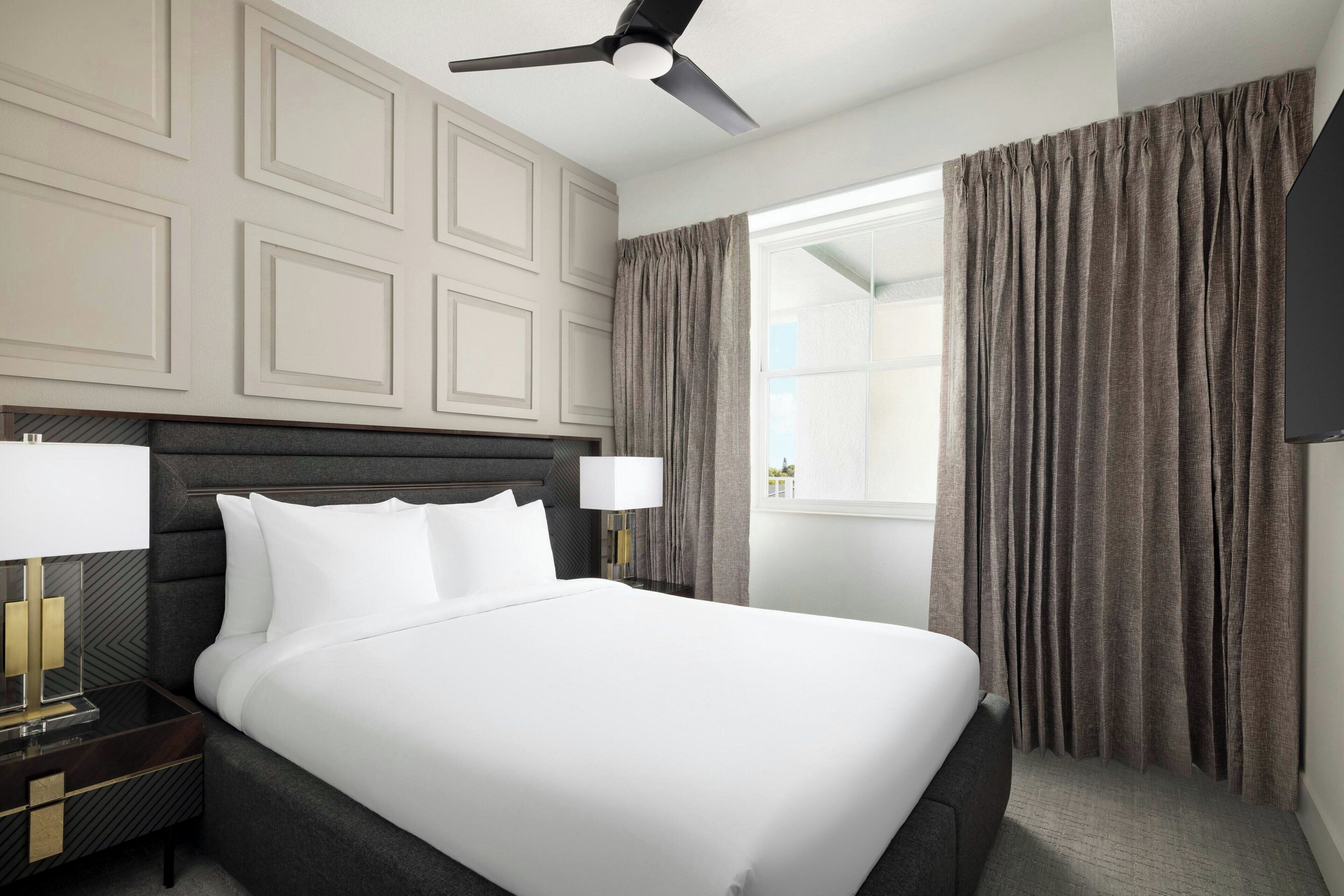 A hotel room with a large bed, a ceiling fan, a nightstand with a lamp, and a view of the outdoors through a window with curtains.